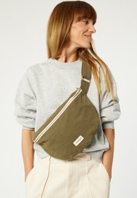 Sac à bandoulière vert olive en tissu, doté d'une fermeture éclair et d'une étiquette décorative. Porté en travers du corps, associé à un sweat-shirt gris.