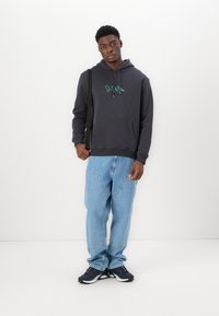 Kapuzensweatshirt in Dunkelgrau mit grünem Grafikdruck, Fronttasche, kombiniert mit hellblauen Jeans und schwarzen Sportschuhen.