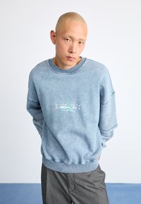 Heathered blå hettegenser med rund hals, senkede skuldre og ribbede mansjetter. Har et grafisk logo i hvitt og lysegrønt foran.