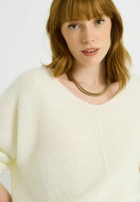 Suéter de punto de gran tamaño en color blanco roto con un amplio escote en V. Textura suave y corte holgado, acompañado de un collar curvado y metálico.