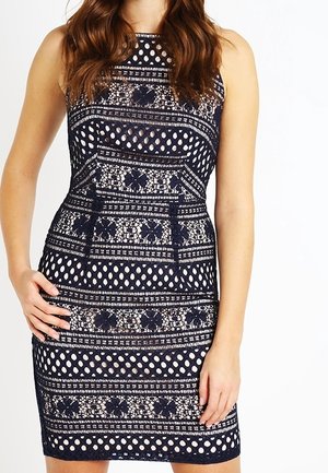 Robe bleu marine sans manches avec des motifs en dentelle complexes et des dessins géométriques, portée par une personne aux longs cheveux bruns.