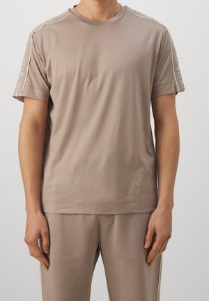 Mężczyzna ubrany w beżowy T-shirt z krótkim rękawem i dopasowane spodnie z markowymi paskami biegnącymi wzdłuż rękawów i boków spodni.