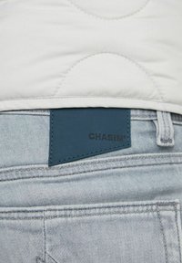 Ljusblå denimjeans med en svart rektangulär läderlapp märkta "CHASIN'" på midjan under en vit vadderad jacka.