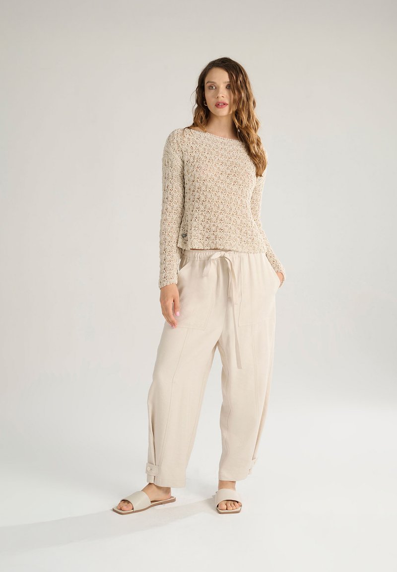 Beige gehäkelter Pullover mit langen Ärmeln, kombiniert mit locker sitzenden, hellbeigen Hose und flachen Sandalen. Weicher Stoff und lässiges Design.