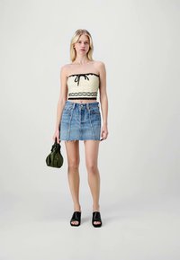 Junge Frau in einem cremefarbenen Tube-Top, blauer Jeans-Minirock, schwarzen offenen Mules, die eine kleine grüne Handtasche hält, vor einem schlichten Hintergrund stehend.