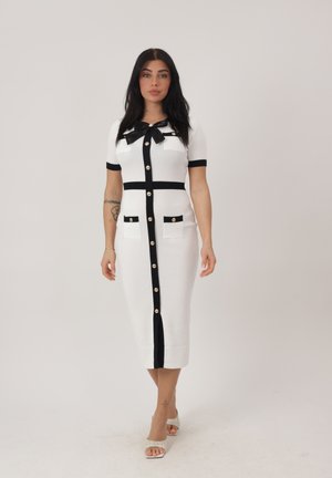 Donna con capelli lunghi e scuri che indossa un abito midi bianco con rifiniture nere, bottoni dorati, dettaglio a fiocco e sandali intrecciati con punta aperta, in piedi davanti a uno sfondo semplice.