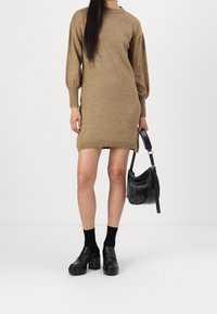 Robe marron en tricot avec un col arrondi et des manches ballon, accompagnée de chaussures plateforme noires et d'un sac à bandoulière noir.