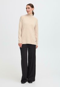 Bežs adīts turtlenecks džemperis, kas kombinēts ar melnām plašām biksēm. Džemperim ir viegls logo elements apakšmalā.