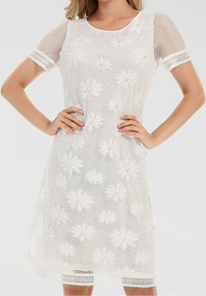 Robe en dentelle blanche avec broderies florales et manches courtes en maille, portée par une personne les mains sur les hanches, sur un fond uni.