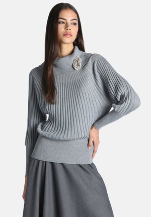 Femme portant un pull gris clair côtelé avec des manches ballon et une broche décorative, associée à une jupe gris foncé.