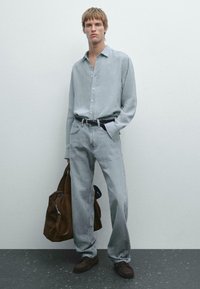 Chemise boutonnée gris clair, jean gris clair décontracté, ceinture noire, chaussures en suedine marron, portant un sac de sport en suedine marron. Style minimaliste.