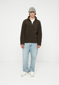 Suéter de forro polar marrón con media cremallera y bolsillo en el pecho, combinado con jeans de mezclilla azul claro y zapatillas blancas. Gorra de béisbol gris.