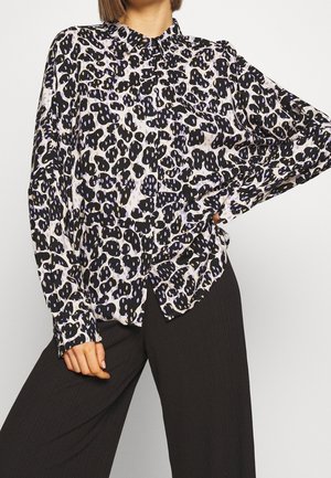 Femme portant une chemise ample à manches longues avec des boutons, ornée de taches léopard noires et blanches, associée à un pantalon noir à jambes larges.