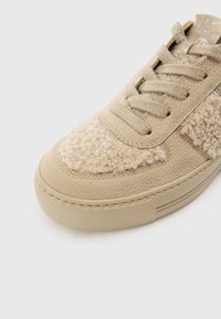 Beige sneaker featuring a textured, fluffy fabric on the toe and a smooth leather body. Flat laces and a thick rubber sole complete the design.
