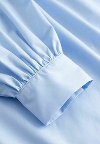 Manche de chemise bleu clair avec tissu froncé et large poignet lisse, mettant en valeur la texture et les détails de la couture.