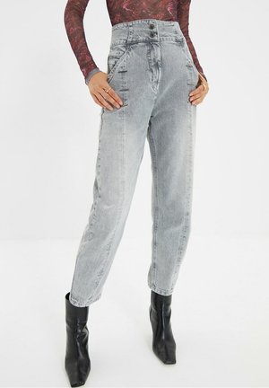 Vrouw met lichtgrijze denim jeans met hoge taille, aangesloten bij de enkels, gecombineerd met zwarte enkellaarsjes met puntige neus en een bordeauxrode top met lange mouwen.