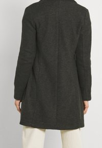 Cappotto di lana verde scuro con taglio dritto, maniche lunghe e colletto. Presenta cuciture minime e una texture liscia.