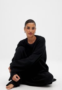 Svart loungekläder-set med en löst sittande sweatshirt och byxor. Mjuk tyg, vida ärmar och avslappnad silhuett. Modell sittande mot en vit bakgrund.