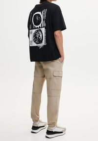 Person, der ein schwarzes T-Shirt mit grafischen kreisförmigen Designs, beigefarbene Cargo-Hosen und weiße Sneaker trägt, steht vor einem weißen Hintergrund.