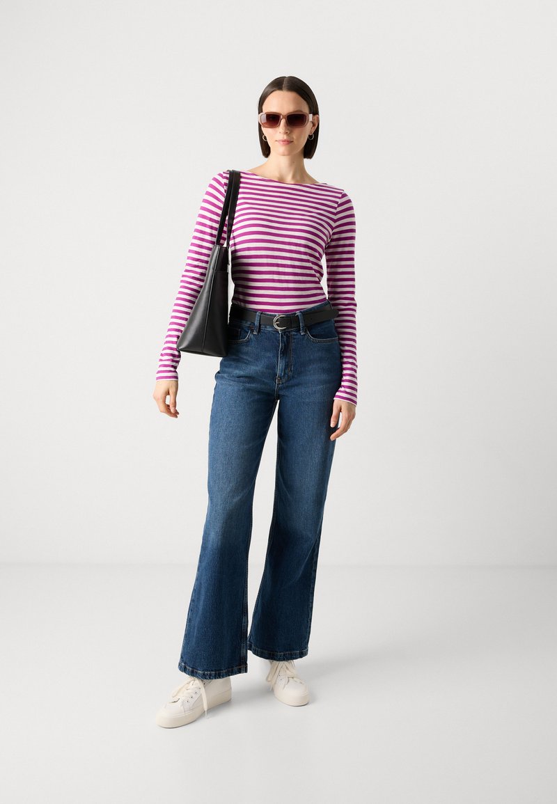 Gestreiftes langärmliges Oberteil in Lila und Weiß, hochgeschnittene blaue Jeans mit Schlag, weiße Sneaker und eine schwarze Tragetasche. Braune Sonnenbrille.