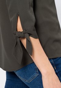 Olivgrünes Hemd mit hochgerollten Ärmeln, das einen Knoten an den Bündchen hat. Der Stoff hat eine subtile Textur und wird mit blauen Denim-Jeans kombiniert.