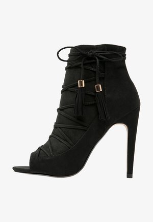 Czarny zamszowy botek z odkrytymi palcami, krzyżującymi się paskami, frędzlami i wysokim szpilkowym obcasem. Gładka faktura i minimalistyczny design.