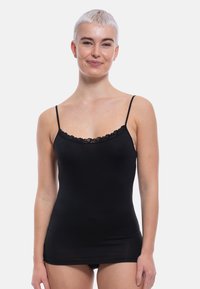 Young woman with short platinum blonde hair wearing a black lace-trimmed camisole top, standing against a plain white background.