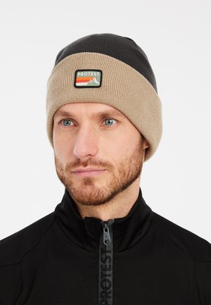 Knit beanie in beige and black, featuring a logo patch with red and green mountain graphics. Soft material with a folded cuff design.