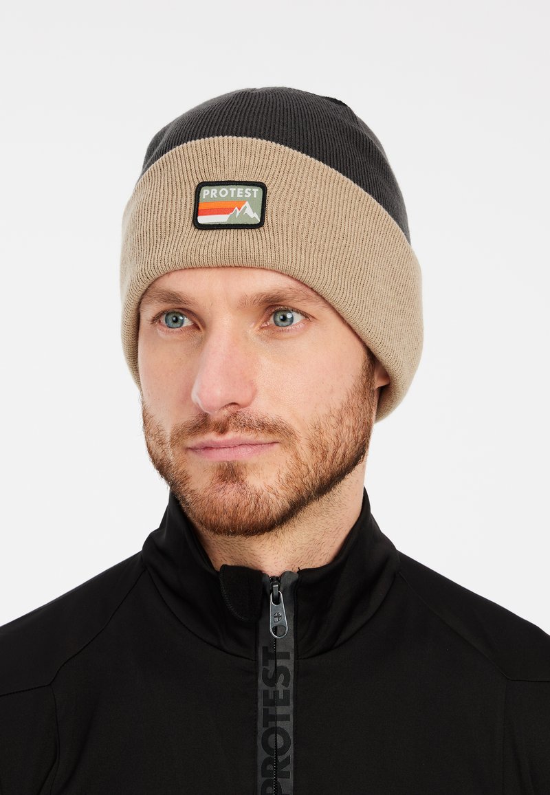 Gebreide beanie in beige en zwart, voorzien van een logopatch met rode en groene berggraphics. Zacht materiaal met een design van omgeslagen rand.