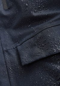 Tessuto resistente all'acqua di colore blu navy con gocce, caratterizzato da una superficie strutturata. Mostra un primo piano delle cuciture e un dettaglio della tasca.