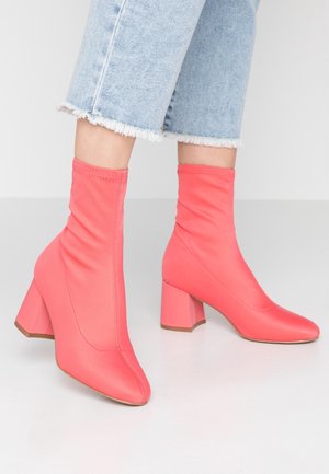 Woman wearing cropped light blue frayed jeans and coral pink mid-calf sock boots with block heels standing on a white floor.
