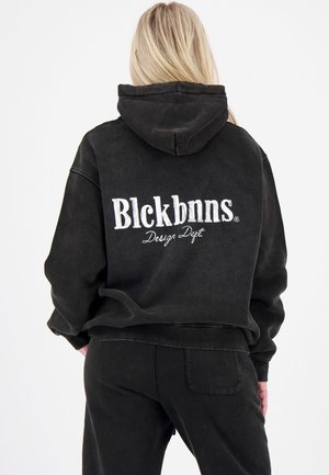 Blonde Person, die einen schwarzen Hoodie und eine Hose trägt, von hinten zu sehen, mit "Blckbnns Design Dept." in weißen Buchstaben auf dem Hoodie.