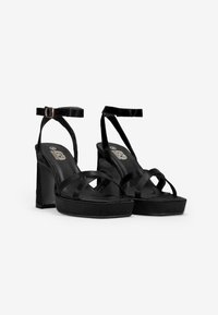 Las sandalias de tacón alto de terciopelo negro cuentan con un diseño de plataforma, tiras entrelazadas y una correa ajustable en el tobillo con una hebilla metálica.