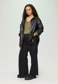Zwarte neplederen overhemdjas, olijfgroene gebreide top met structuur, zwarte brede jeans en zwarte schoenen met dikke zool. Model poseert met één hand in de zak.
