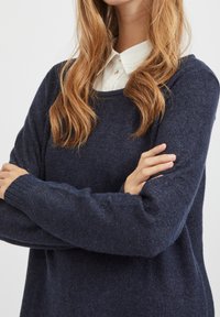 Navyblauer Strickpullover mit langen Ärmeln, rundem Ausschnitt und gerippten Bündchen. Verfügt über eine weiche Textur und eine lässige Passform, die über einem weißen Hemd mit Kragen getragen wird.