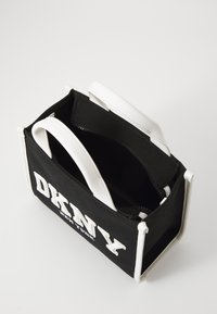 Sac en toile noir avec des accents blancs, doté d'un zip sur le dessus et portant le logo "DKNY" imprimé. Design compact avec deux poignées.