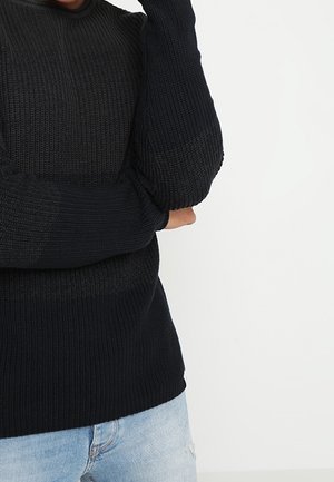 Osoba nosząca czarny dziergany sweter z rękawami częściowo zakrywającymi dłonie, w połączeniu z jasnoniebieskimi jeansami na jednolitym białym tle.