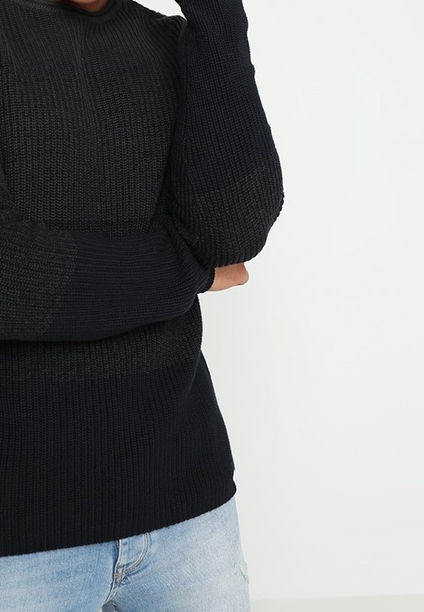 Personne portant un pull en tricot noir avec des manches couvrant partiellement les mains, associé à un jean bleu clair sur un fond blanc uni.