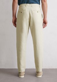 Pantalons en lin beige clair à coupe droite, dotés de deux poches arrière et d'accents de boutons dorés. Texture lisse, design minimaliste.