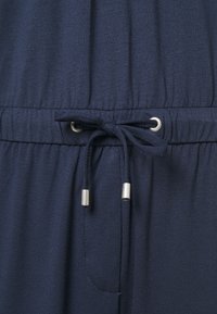 Tejido azul marino con un ligero brillo, que cuenta con una cinturilla fruncida y un cordón con extremos metálicos. Superficie suave y texturizada.