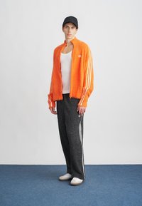 Chaqueta de felpa naranja con cremallera y rayas blancas, camiseta sin mangas blanca, pantalones negros texturizados con acentos blancos y zapatillas blancas. Gorra negra.