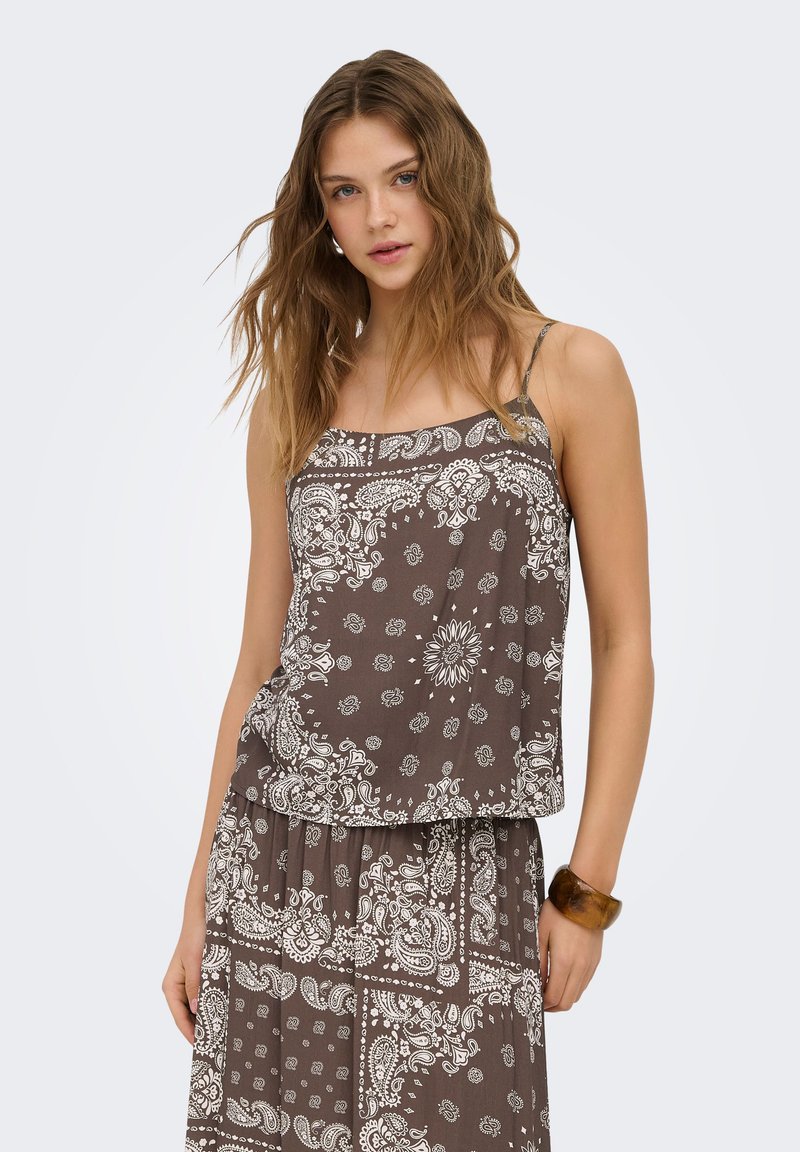 Jeune femme aux cheveux ondulés portant un haut sans manches marron à motif paisley et une jupe assortie, debout avec une main dans la poche.