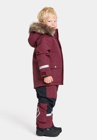 Burgundy vinterjacka med huvtröja i päls, grå reflexdetaljer, kombinerad med mörka byxor och svarta stövlar, designad för kallt väder.