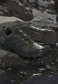 Chaussures de randonnée Terrex noires et gris foncé avec semelles texturées et passants jaunes, posées sur un sol rocheux humide près de grosses pierres.