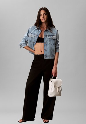 Vrouw met cropped spijkerjack, zwarte bralette, wijde zwarte broek, houdt een witte rugzak vast, staat tegen een effen grijze achtergrond.