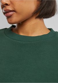 Felpa verde scuro con scollatura a girocollo a coste. Il tessuto appare morbido, con una consistenza liscia e senza motivi o dettagli visibili.