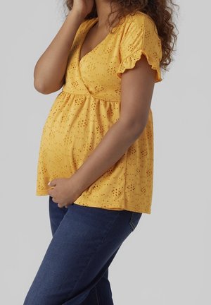 Femme enceinte tenant son ventre d'une main, portant un chemisier jaune à manches courtes en œillets et un jean bleu, sur un fond gris.