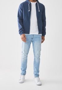 Námořnická zipová mikina s šňůrkami, bílý tričko s kulatým výstřihem, světle modré slim-fit džíny a bílé tenisky. Neformální, pohodlný outfit.