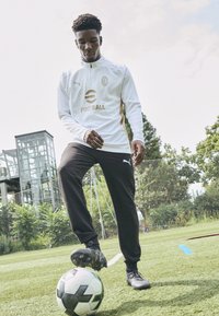 Weißes langärmliges Sportshirt mit goldenen Akzenten, kombiniert mit schwarzen Jogginghosen. Schwarze Fußballschuhe auf einem Fußball auf grünem Rasen.