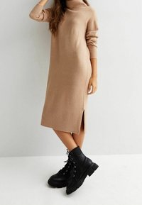 Robe pull en tricot beige clair avec col roulé, ourlet côtelé et fente latérale ; associée à des bottines noires épaisses à lacets.
